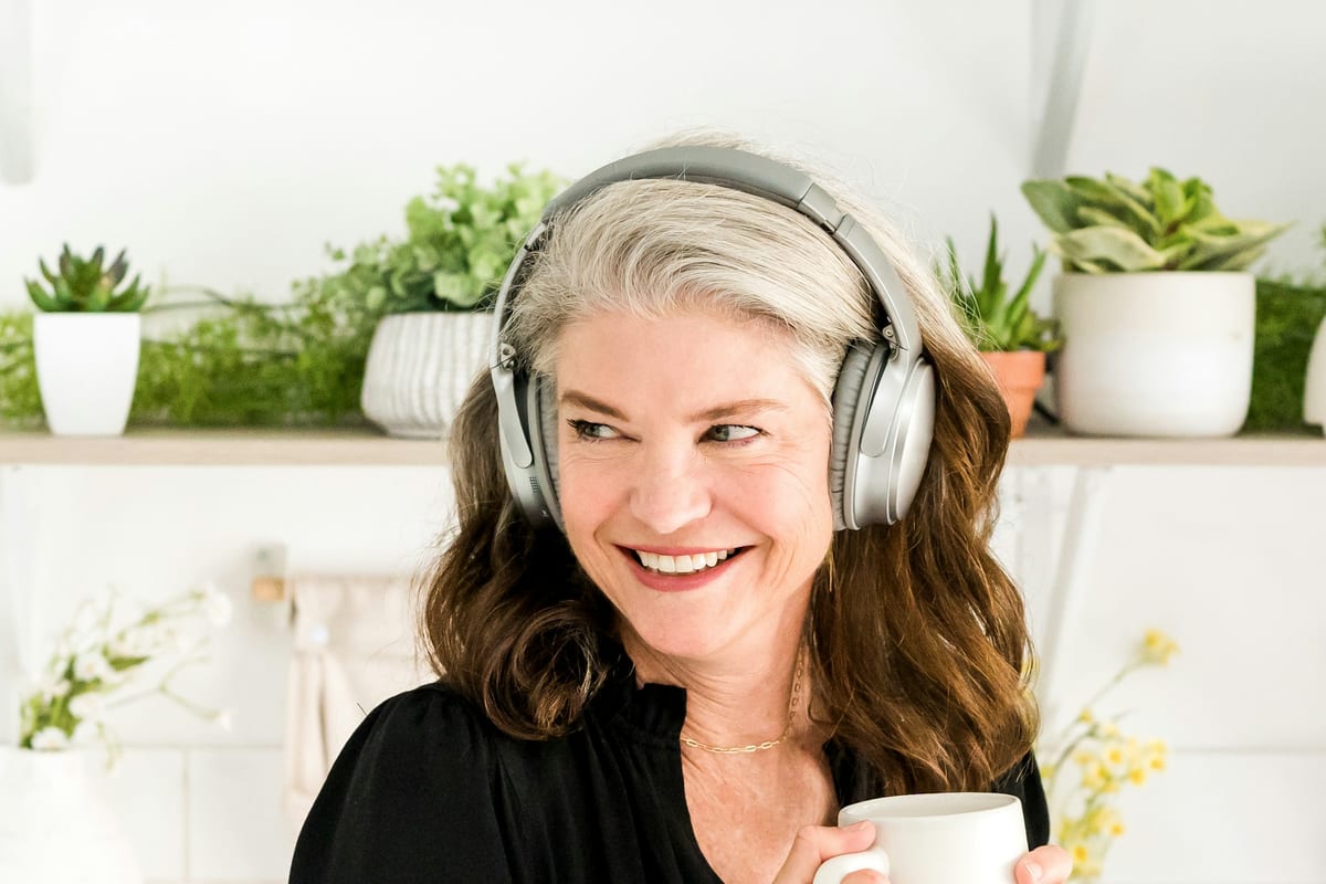 Una mujer sonriendo con auriculares de diadema y una taza en la mano, en una cocina con plantas