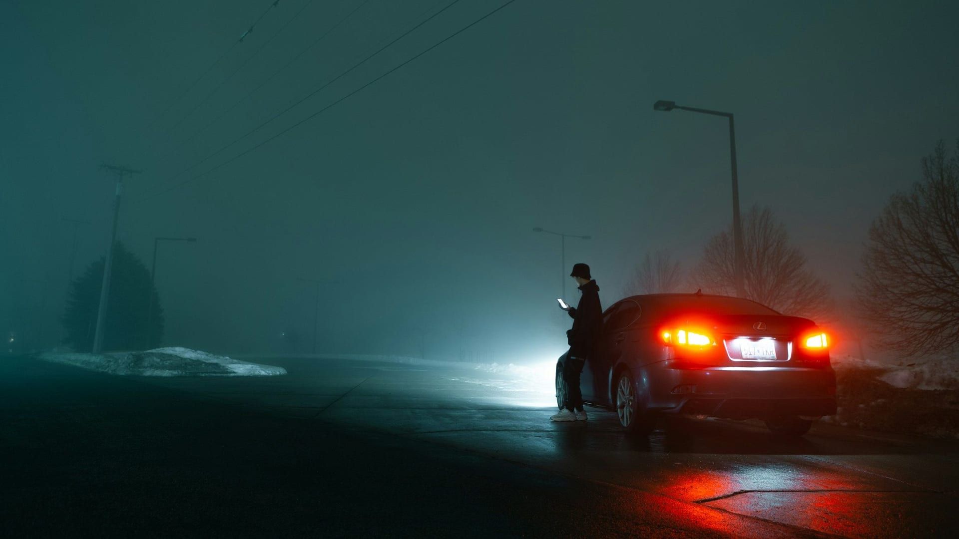 Un coche parado de noche en una carretera con niebla, con las luces encendidas y su conductor de pie al lado