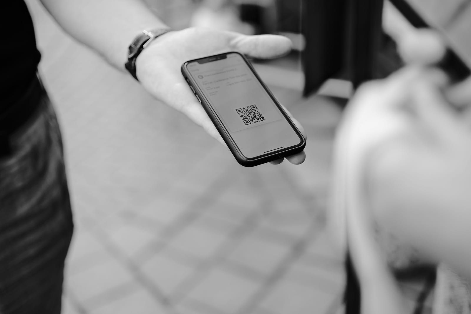 En blanco y negro, una mano enseña a otra persona un móvil con un código QR en la pantalla