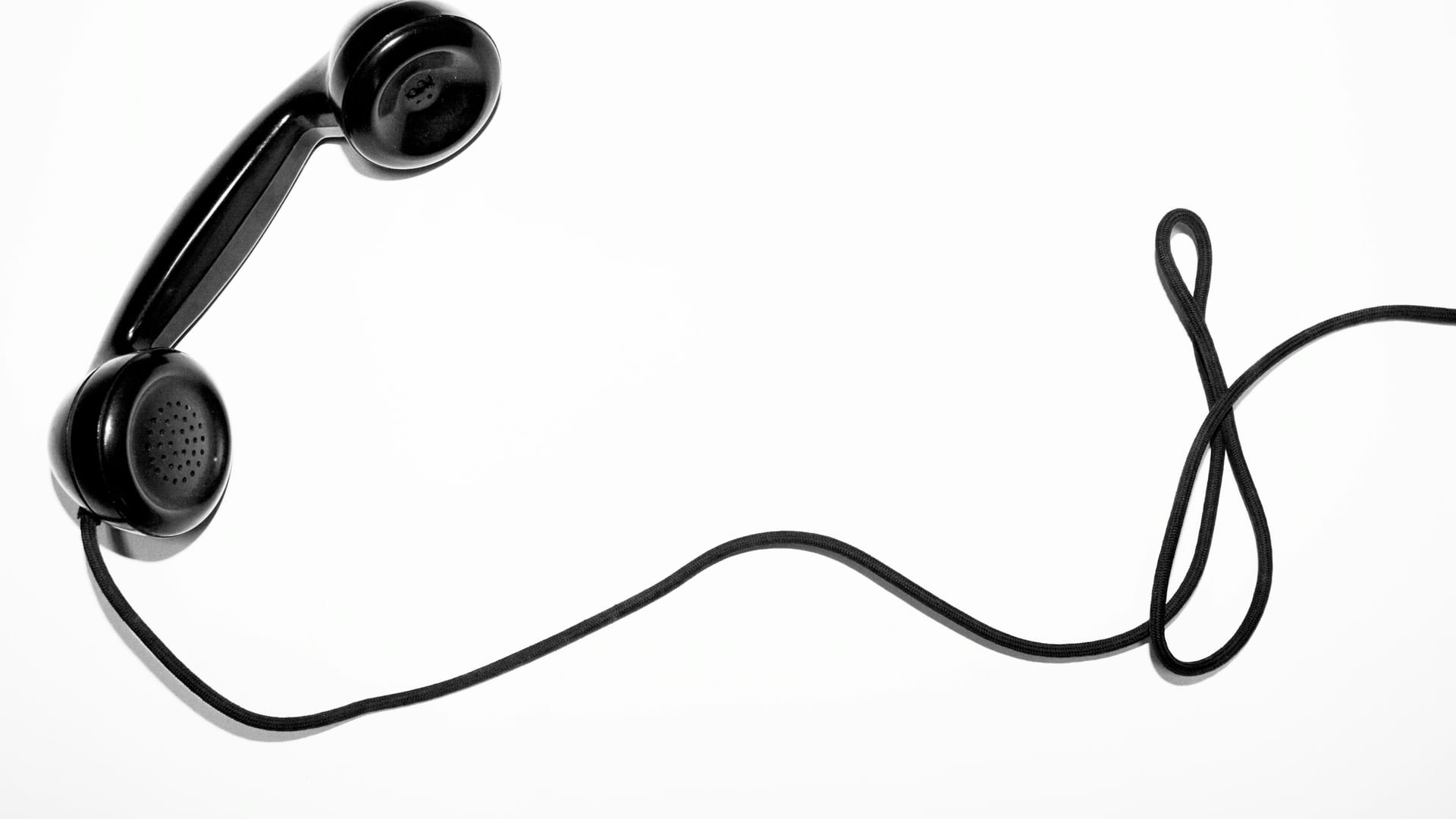 Un auricular de teléfono negro descolgado sobre fondo blanco, con el cable en el aire