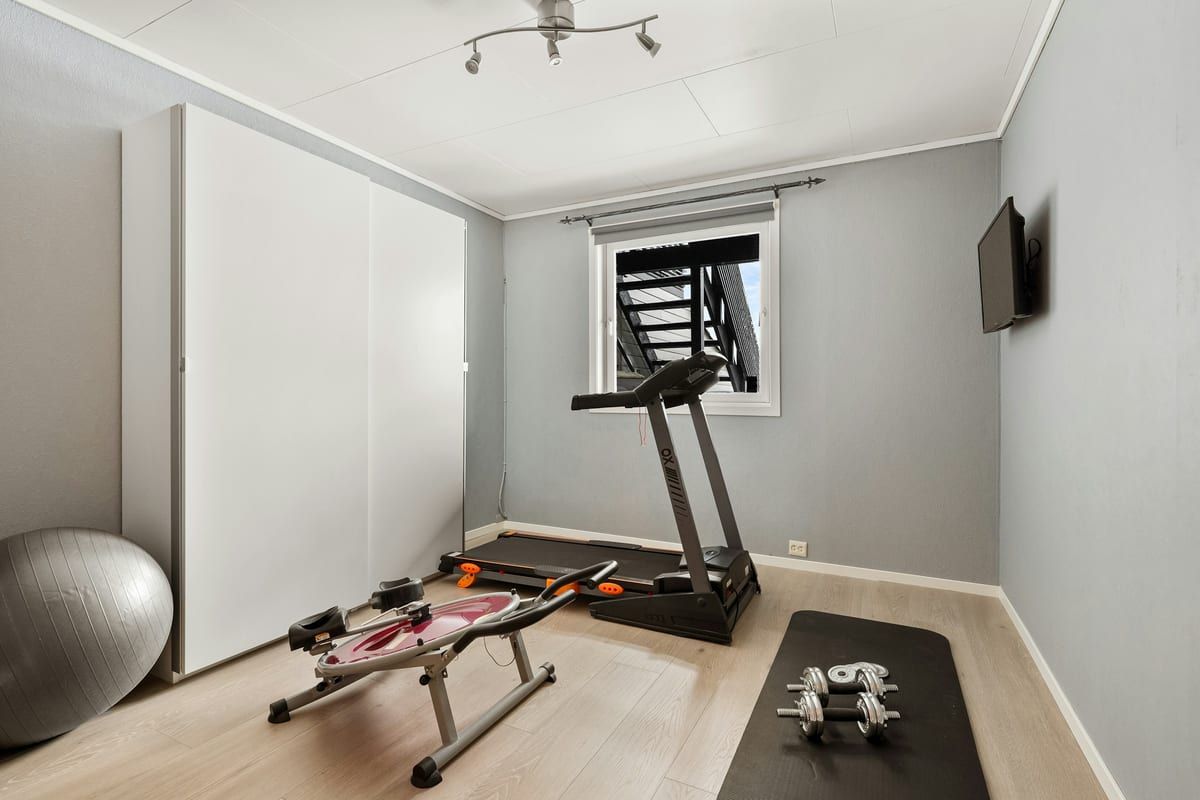 Cinta de correr junto a una ventana en un cuarto de casa con una tele en la pared y aparatos de gimnasio