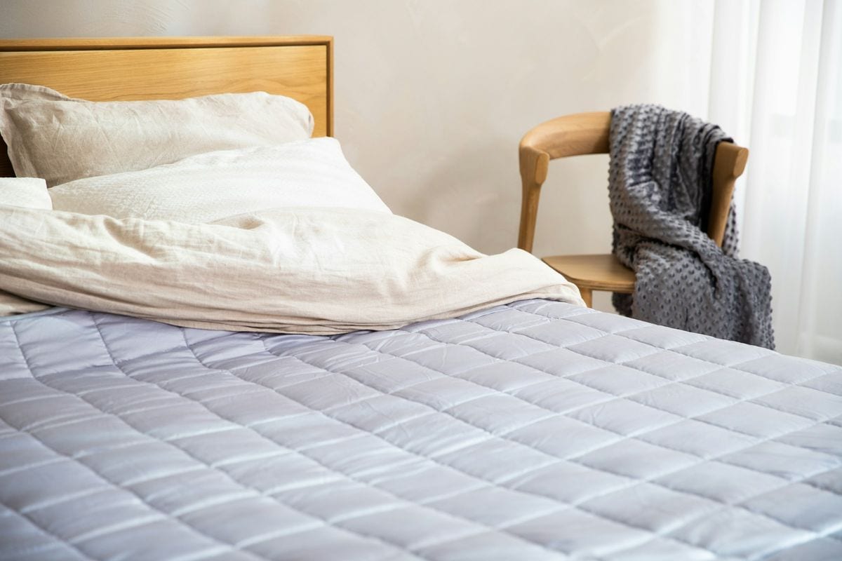 Cama con una colcha acolchada gris claro, almohadas blancas y una manta de punto gris sobre una silla de madera