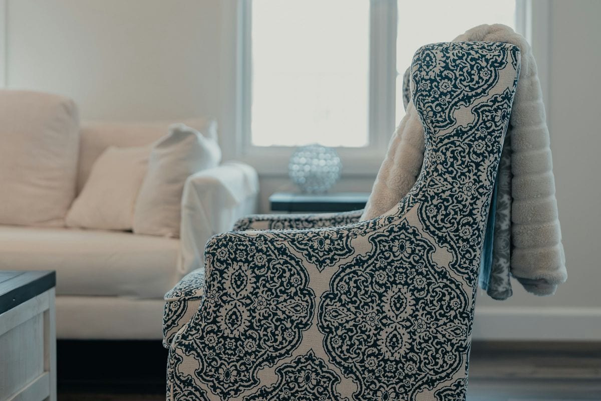 Sillón tapizado con estampado azul y una manta de pelo beis colgada del respaldo, junto a una ventana