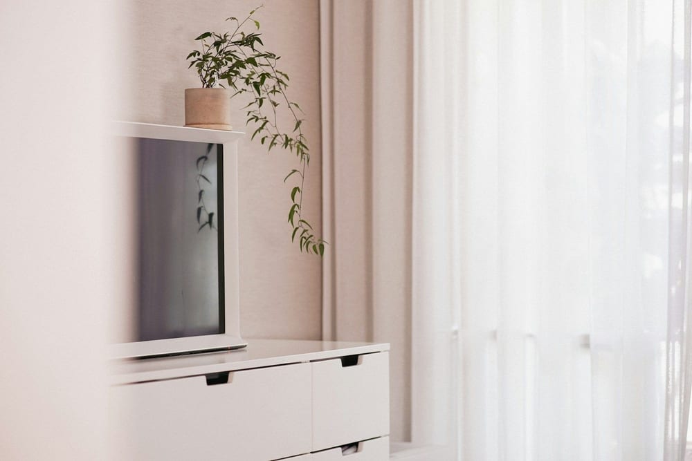Televisor apagado encima de una cómoda blanca, junto a una ventana con cortinas y una planta