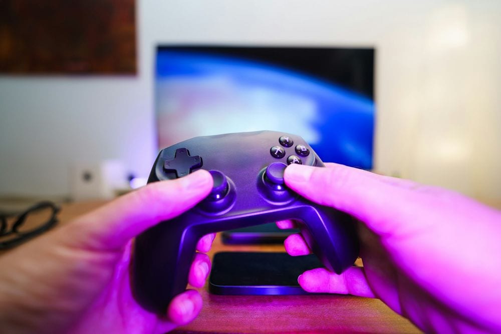 Manos con un mando de consola delante de una tele encendida en una habitación con luz morada