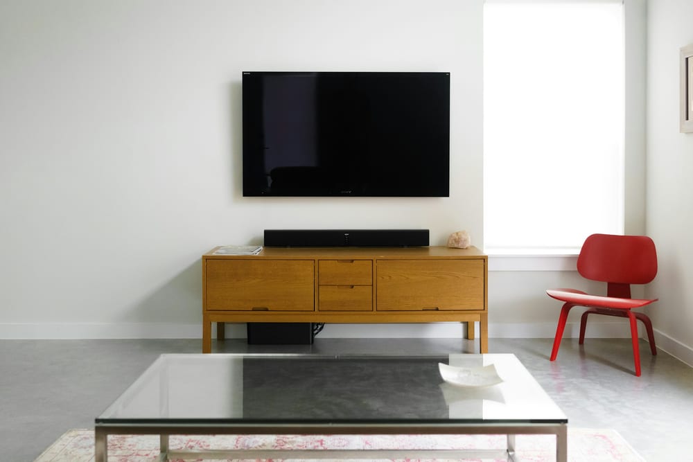 Televisor colgado en una pared blanca sobre un mueble de madera con una barra de sonido, y una silla roja al lado