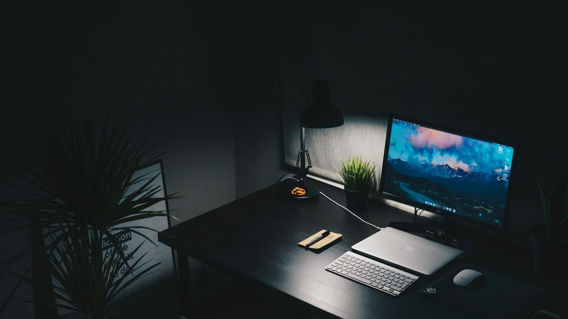 Un escritorio de noche con un monitor encendido, un teclado y una lámpara que ilumina la mesa