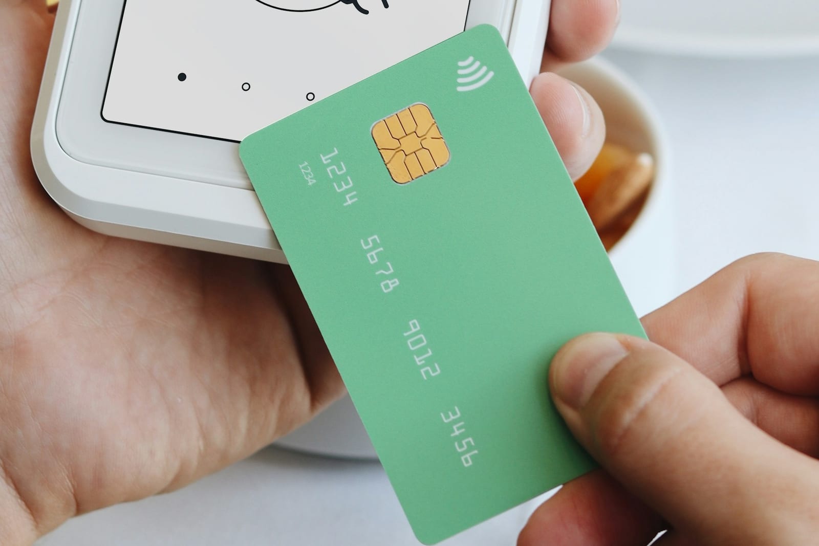 Una mano sostiene una tarjeta bancaria verde junto a un terminal de pago