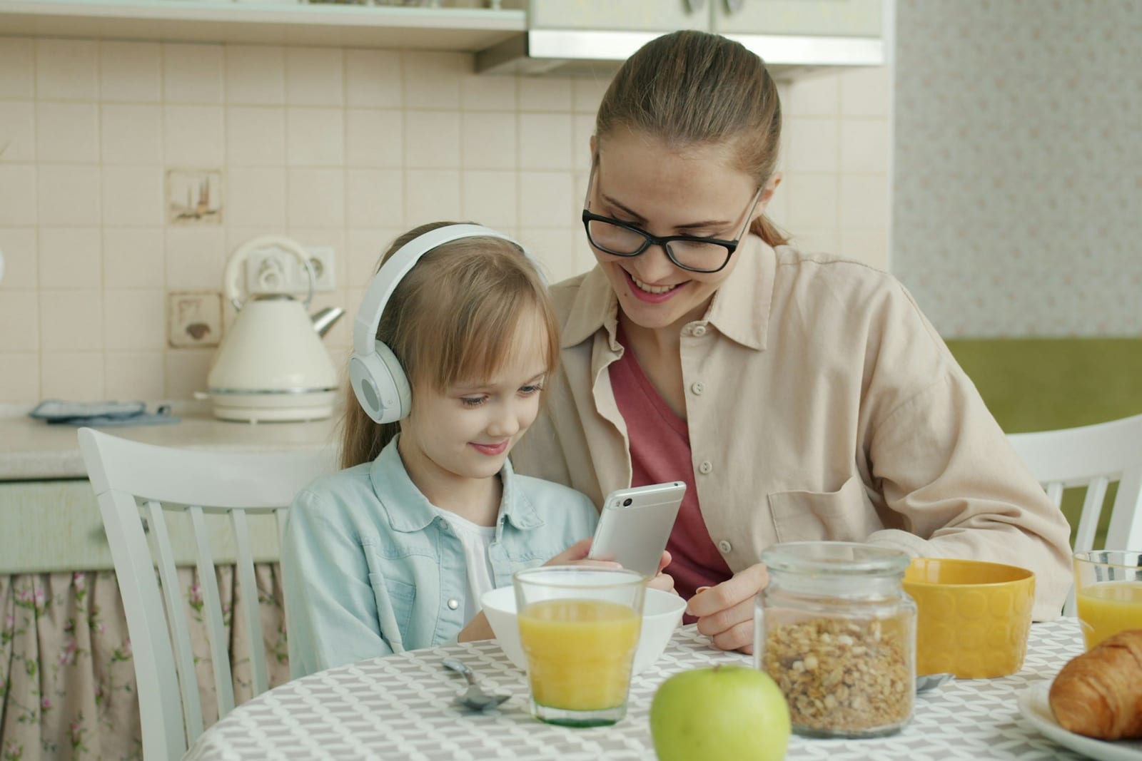Una madre y su hija desayunan en la cocina, la niña con auriculares y la madre con el móvil