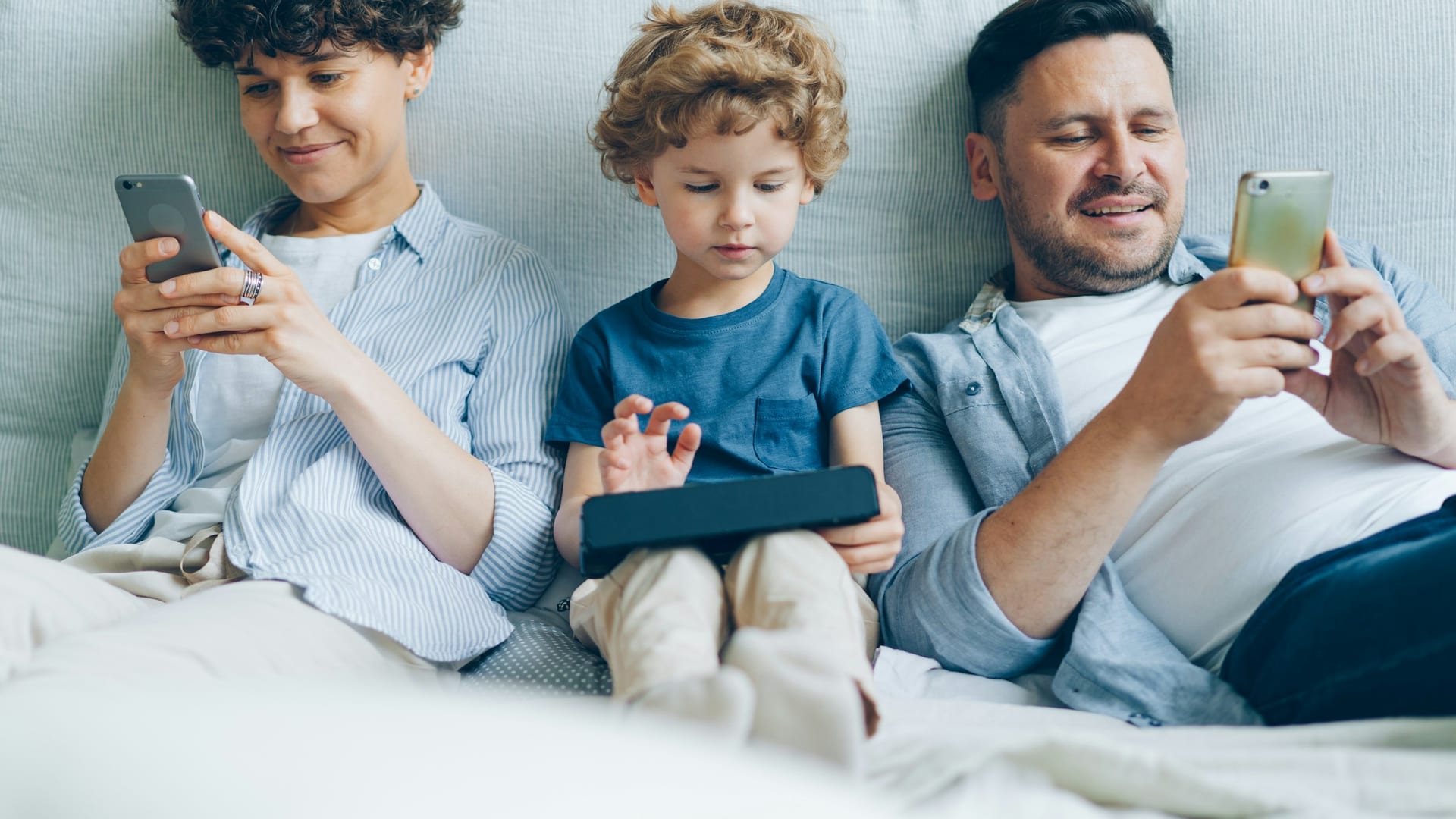 Una madre, un padre y su hijo en el sofá, cada uno mirando su móvil o su tableta